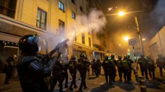 A riot policeman fires tear gas over the heads of a line of riot police ahead of him, in a nighttime urban environment.