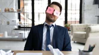 A man in a business suit sits at his desk, asleep but with Post-It notes on his eyes with eyes drawn on.