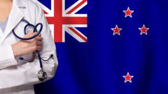 A close-up of a doctor with a stethoscope against a backdrop of the New Zealand flag.