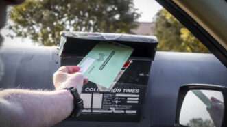 A hand extends from a car and places a ballot in a mail box.