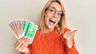Smiling blonde women gives a thumbs up and holds a handful of Norwegian kroner, fanned out.