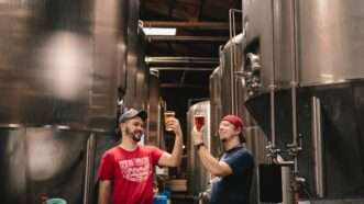 Two men holding full beer glasses up amid tall stainless steel tanks in a brewery.
