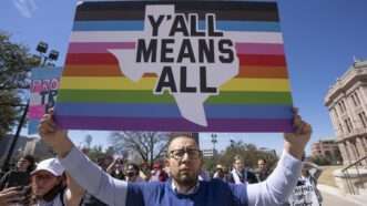 Person protesting in Texas with rainbow striped sign that says "Y'all means All"